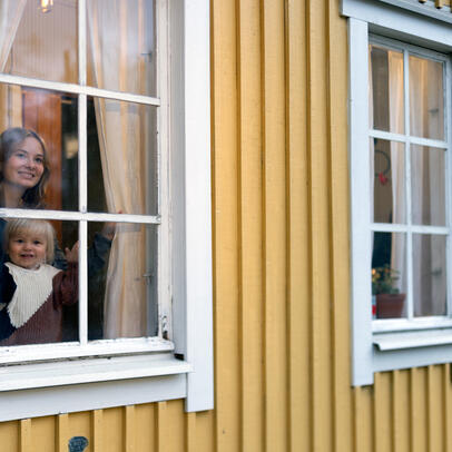 Pieni hymyilevä tyttö ja nainen kurkistaa puutalon ikkunasta.