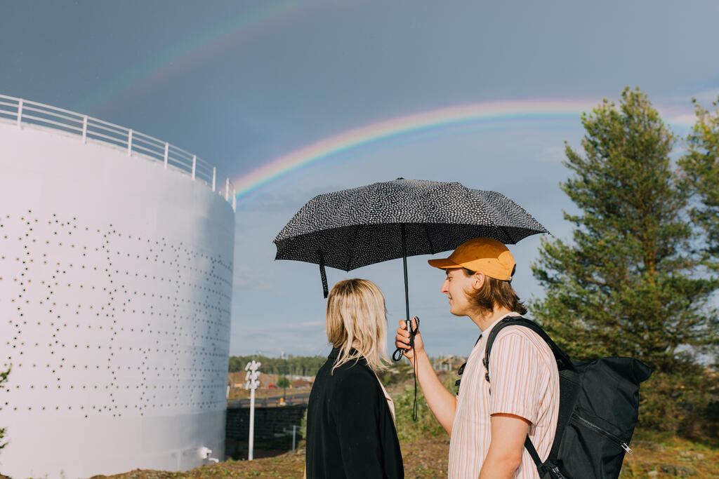 Kaksi henkilöä sateenvarjon alla Kruunuvuorenrannassa, taustalla sateenkaari ja säiliö.