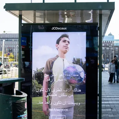 Helsinkiläinen bussipysäkki, jossa on arabiankielinen 700 000 helsinkiläistä -kampanjan mainos.