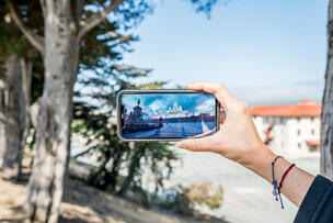 : A hand holding a phone, with the phone's camera reflecting a cityscape.