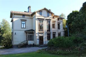 En ljus trävilla i tre våningar som befinner sig i skuggan av grönskande träd.
