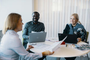 Kolme henkilö istuu tietokoneiden ääressä ja juttelee.