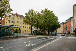 There are cars and a tram on Mäkelänkatu. In the background, there are apartment buildings, and the leaves on the trees are starting to turn yellow.
