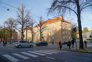 Risteys, jossa etualalla suojatie ja jalankulkijoita, taustalla risteyksessä ajavia autoja ja taustalla kivinen kerrostalo.