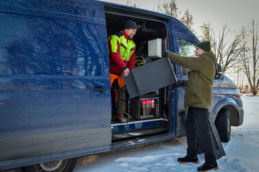 Kaksi miestä nostaa mustaa pönttöä sinisestä pakettiautosta.