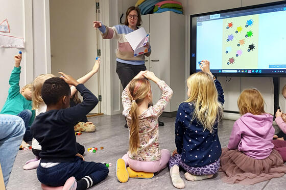 Lapset istuvat lattialla opettajan ja ison näyttöruudun edessä.