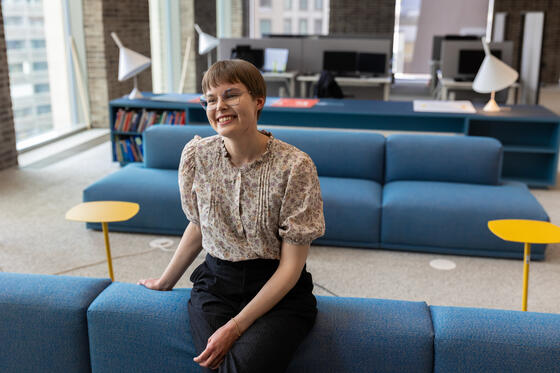 Maisema-arkkitehti Sofia Kangas nojaa hymyillen sohvaan työpaikkansa suuressa toimistohuoneessa, jossa on moderneja huonekaluja ja valaisimia. 