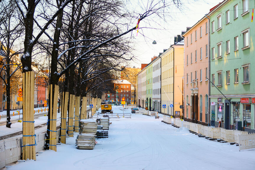 Kuva tyhjästä lumisesta kadusta. Kadun reunustoilla on työmaa-aitoja, rakennustarvikkeita ja laudoilla suojattuja katupuita. Reunassa näkyy värikkäitä kerrostaloja.