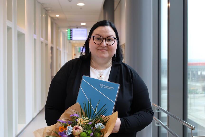 En person står i en ljus korridor och håller en blombukett och en blå mapp med en organisations logotyp. I bakgrunden syns korridoren och utsikten genom fönstren.