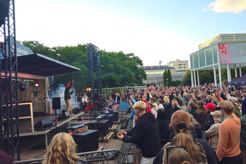 Yleisö on kerääntynyt lavan eteen Ala-Malmin puistossa. Taustalla näkyy Malmitalo ja paviljonki.
