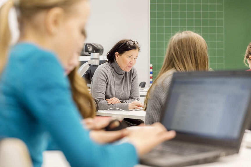 Oppilas tietokoneella luokassa, taustalla opettaja