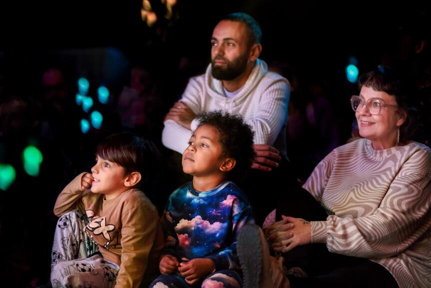 Två barn och två vuxna följer föreställningen i en sal med dimljus.