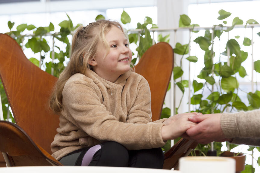 Tyttö istuu oranssissa nojatuolissa ja pitää aikuista kädestä.