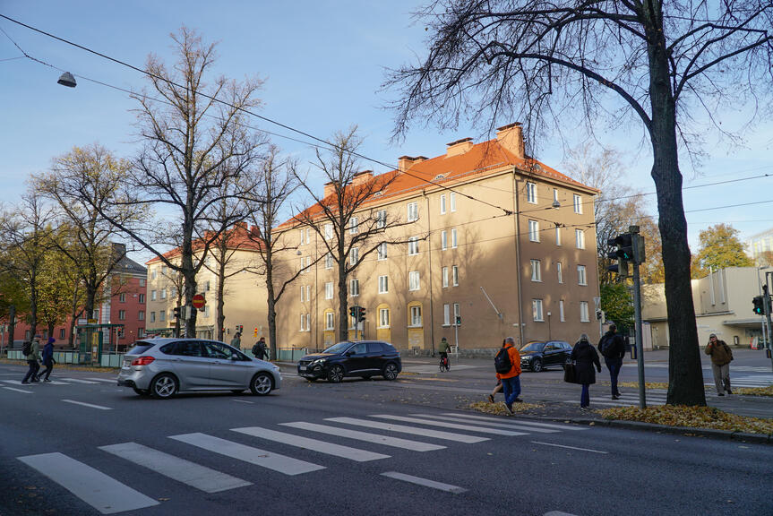 Risteys, jossa etualalla suojatie ja jalankulkijoita, taustalla risteyksessä ajavia autoja ja taustalla kivinen kerrostalo.