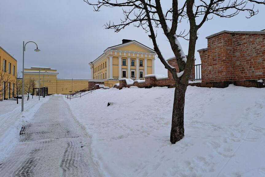 Talvinen maisema, jossa luminen kävelytie kulkee vasemmalla ja oikealla puolella näkyy paljas puu sekä punatiilinen muuri. Taustalla kohoaa vaaleankeltainen, klassistinen rakennus.