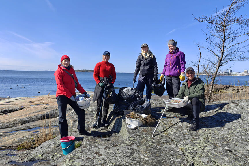 Ihmisiä roskansiivousvälineiden kanssa. Taustalla näkyy sininen taivas ja meri.