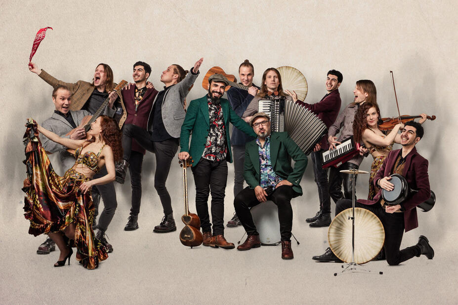 Thirteen exuberant musicians stand side by side against a beige background, some holding instruments.