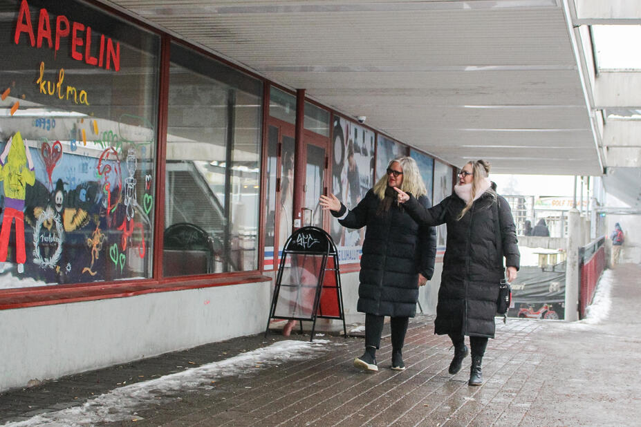 Two women wearing black padded jackets are walking in front of a colorful window.