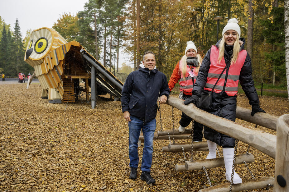 Mies ja kaksi naista syksyisessä puistossa. Takana pöllön näköinen liukumäki