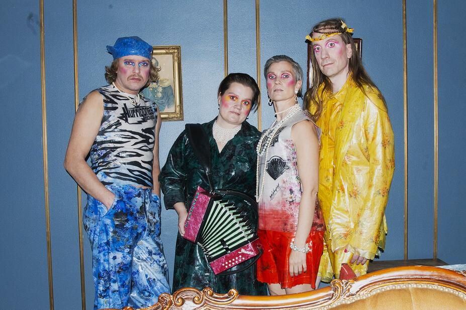 Four distinctively dressed people stand in front of a blue wall, looking at the camera.