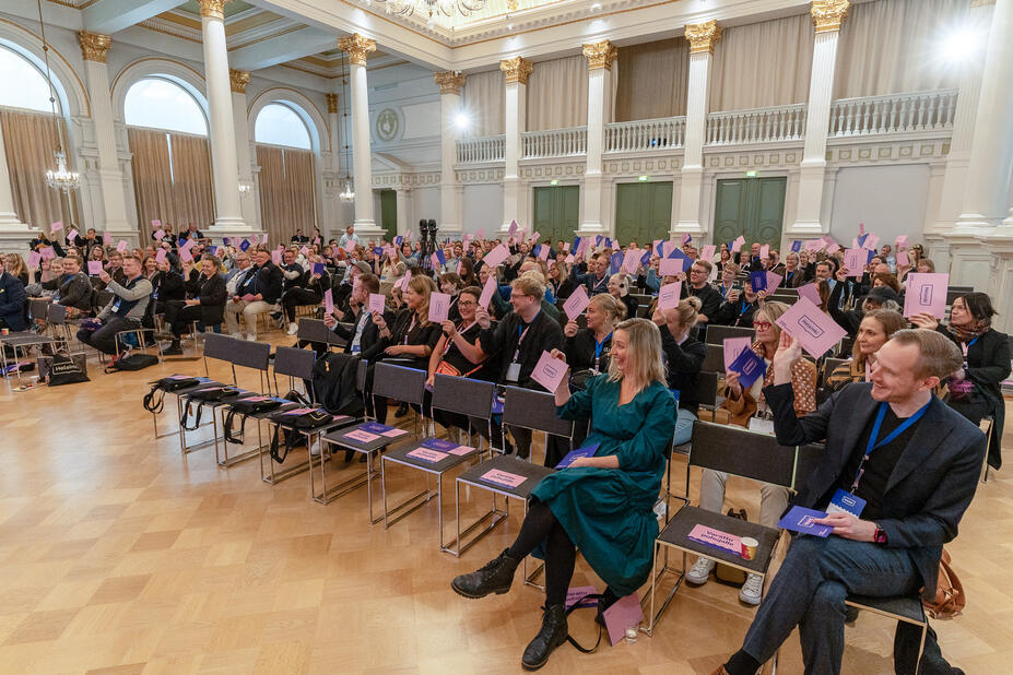 Yleisö istuu salissa ja vilkuttaa vaaleanpunaisia lappuja hymyillen.