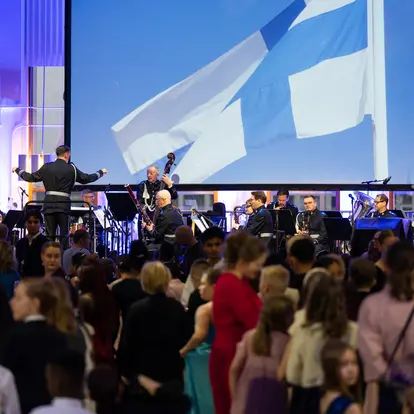 Children dancing with the Finnish flag and an orchestra in the background.