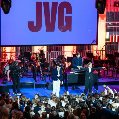 JVG performs on stage with a group of children in front of the stage.