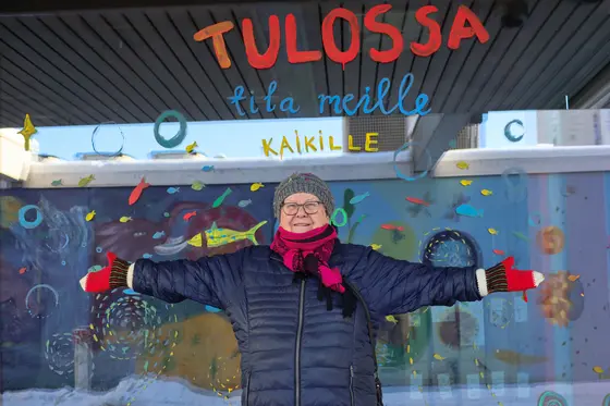 Liisa Aunula med armarna öppna åt sidorna framför ett färgglatt fönster.