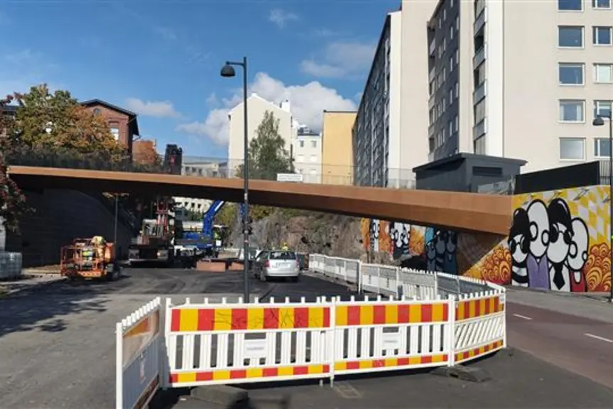A rust-colored bridge over the street. There is roadwork on the street, with cliffs, apartment buildings, and trees along the edge.