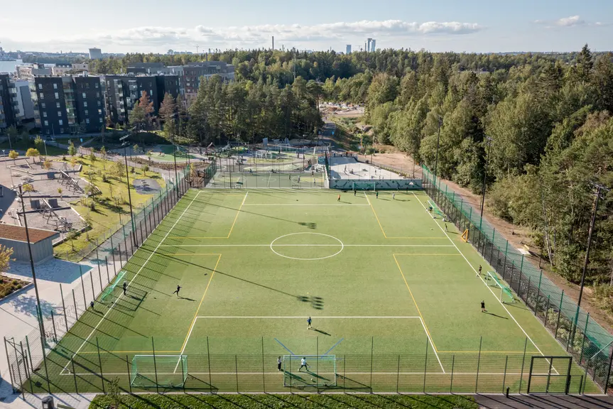 Ilmakuva tekonurmikentällä ihmisiä pelaamassa jalkapalloa. Taustalla näkyy kerrostaloja ja metsää.