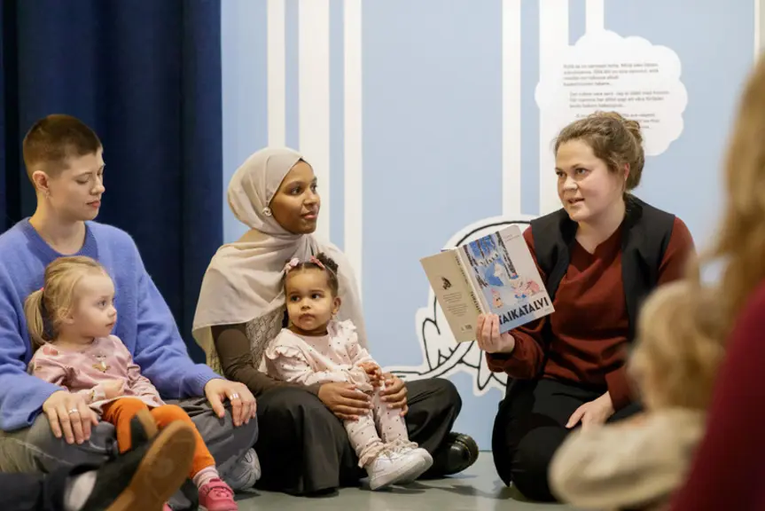 Föräldrar med barn sitter på golvet och lyssnar när man läser en Muminbok för dem.