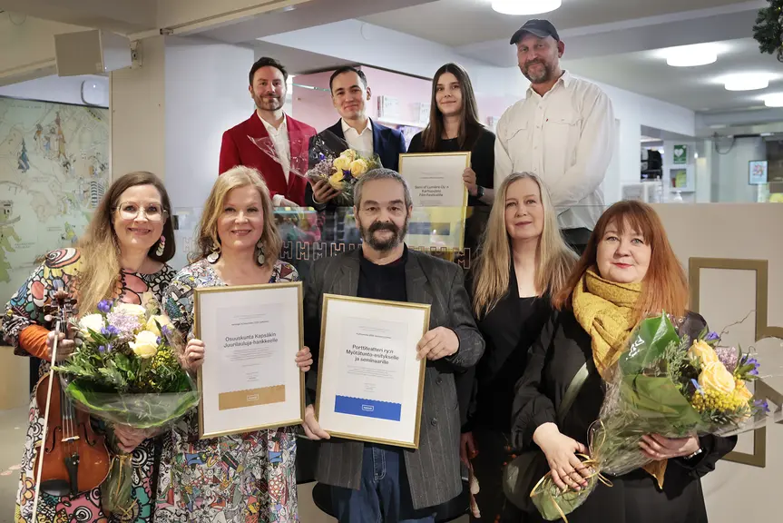 A group of award-winning individuals smile at the camera, holding flowers and certificates in their hands.