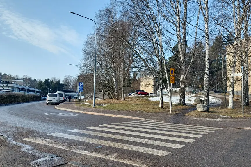 Oikealle kaartuva katu, etualalla on suojatie. Vasemmalla näkyy matala rakennus ja oikealla korkeita koivuja. Taivas on selkeä.