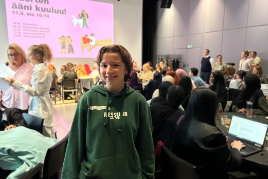 Nuori katsoo kameraan hymyillen isossa salissa, taustalla ihmisiä istuu pöydissä. Takaseinälle on heijastettu esitys, jossa lukee Nuorten ääni kuuluu. 