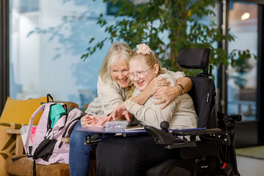Nainen sohvalla ja tyttö pyörätuolissa halaavat