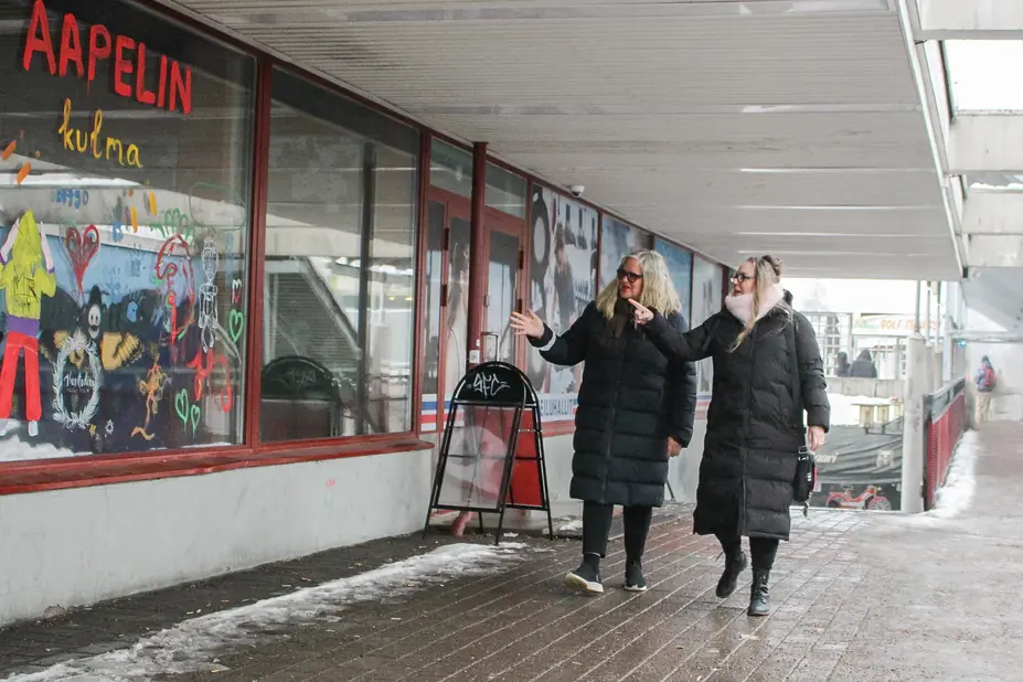 Två kvinnor i svarta vadderade jackor går framför ett färgglatt fönster.