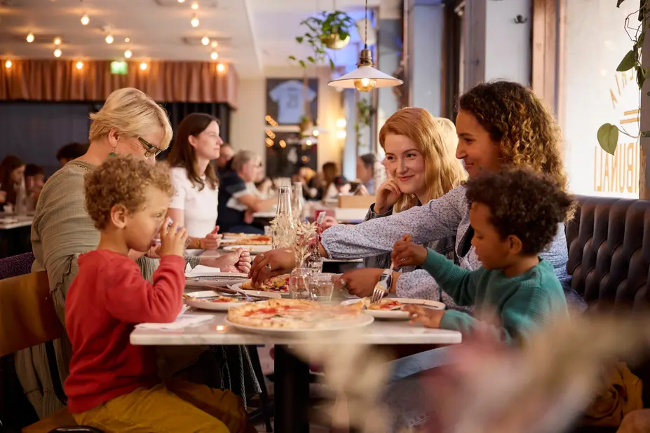 Joukko ihmisiä lapsesta aikuiseen syö pizzaa ravintolan pöydässä.