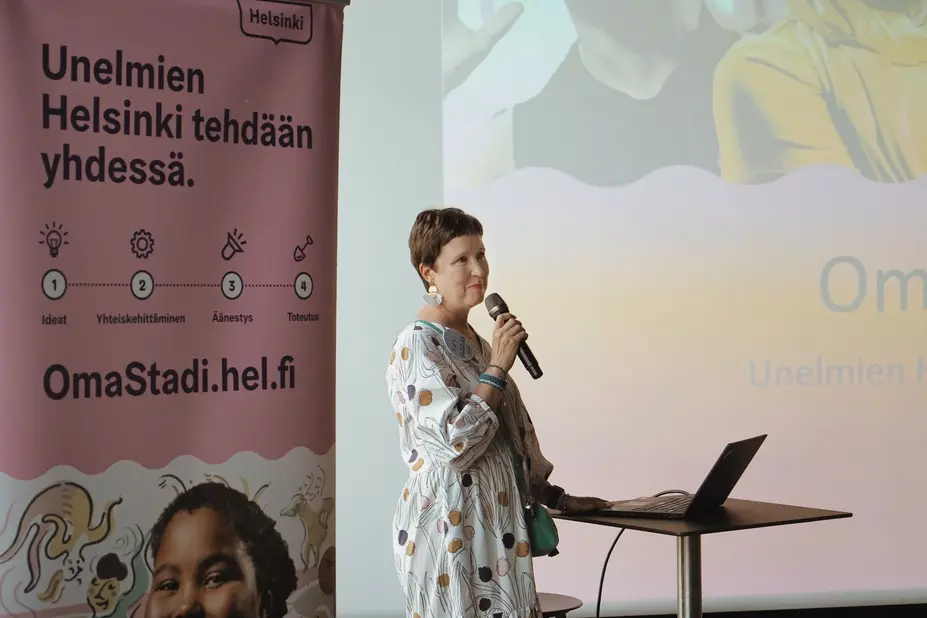 Kirsi Verkka puhuu mikrofoniin valkokankaan edustalla.