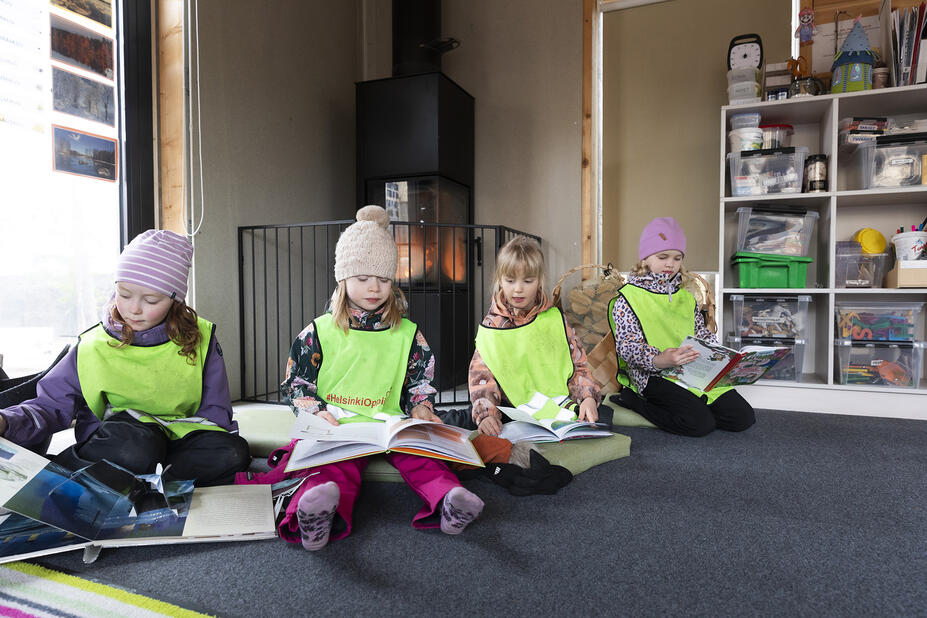 Neljä lasta istuu ulkovaatteissa kodan lattialla ja lukee kirjoja