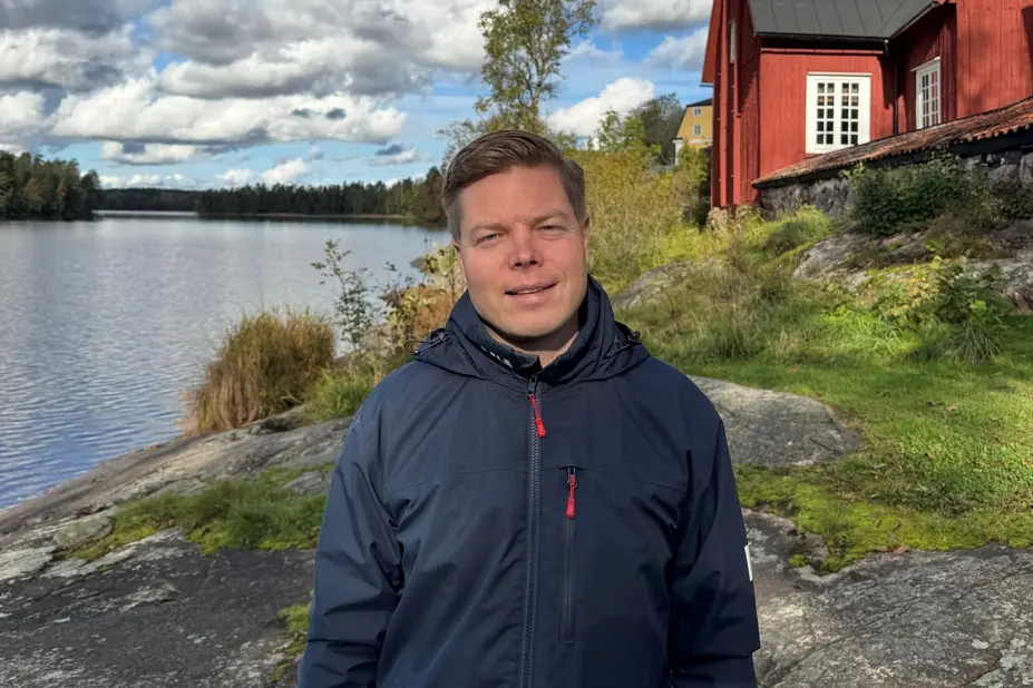 Kai Knocke by the sea, with a red, wooden building in the background. 