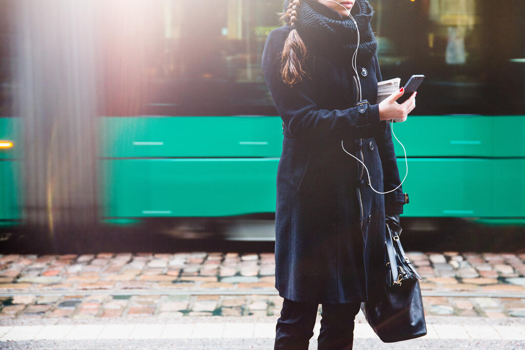 Nainen selaa puhelinta raitiovaunu taustalla. Kuva: Jussi Hellsten