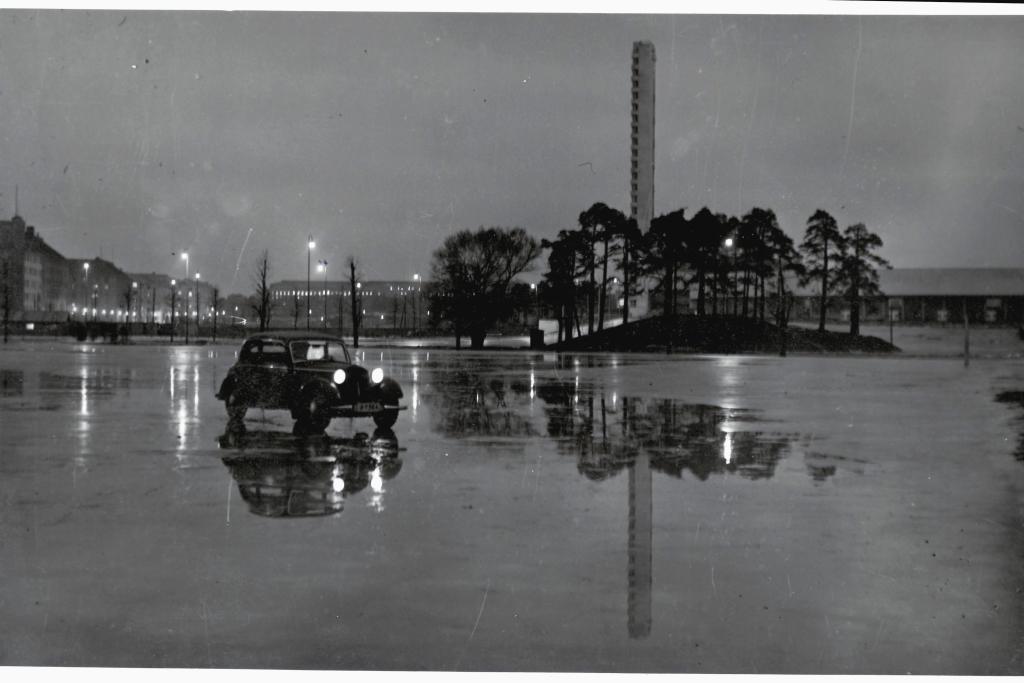 Auto seisoo sateisella kentällä Olympiastadionin tornin edessä. Kuva: Kaupunginarkisto
