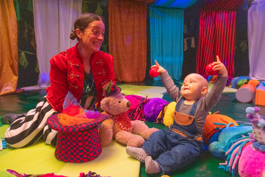 Toddler at the theatre with a circus performer, surrounded by balls and plushies.