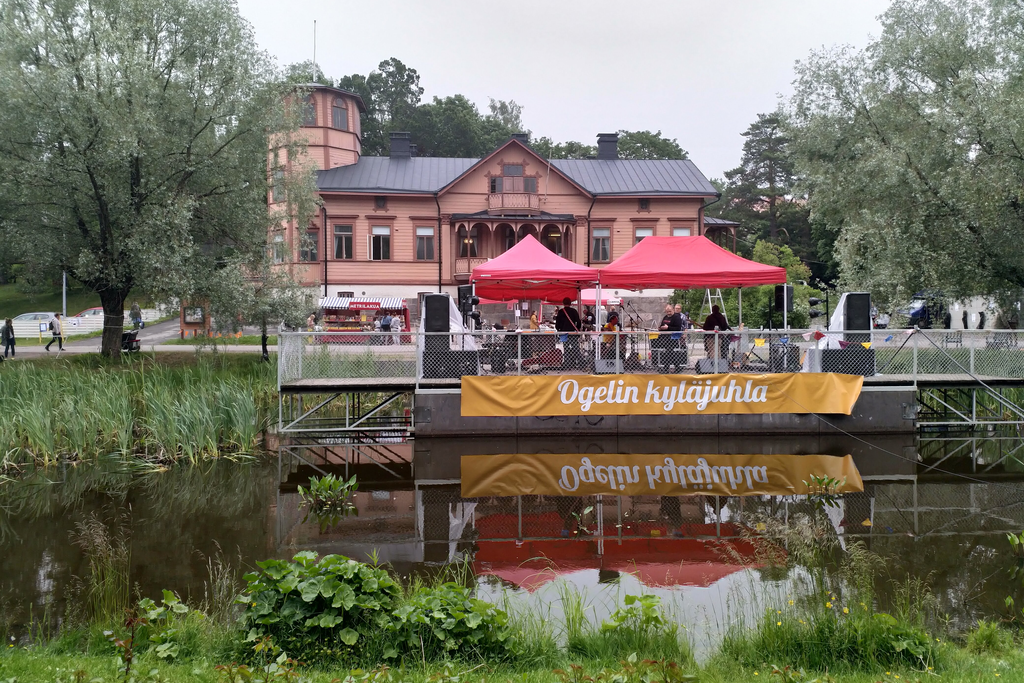 Kuvassa Oulunkylän Seurahuone ja edustalla telttoja ja ihmisiä sekä Ogelin kyläjuhla -lippu.