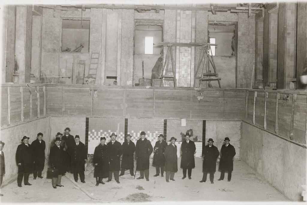 The photo shows 16 men in dark outdoor clothing at the bottom of the unfinished indoor swimming pool. In the background above, a man is plastering a wall.