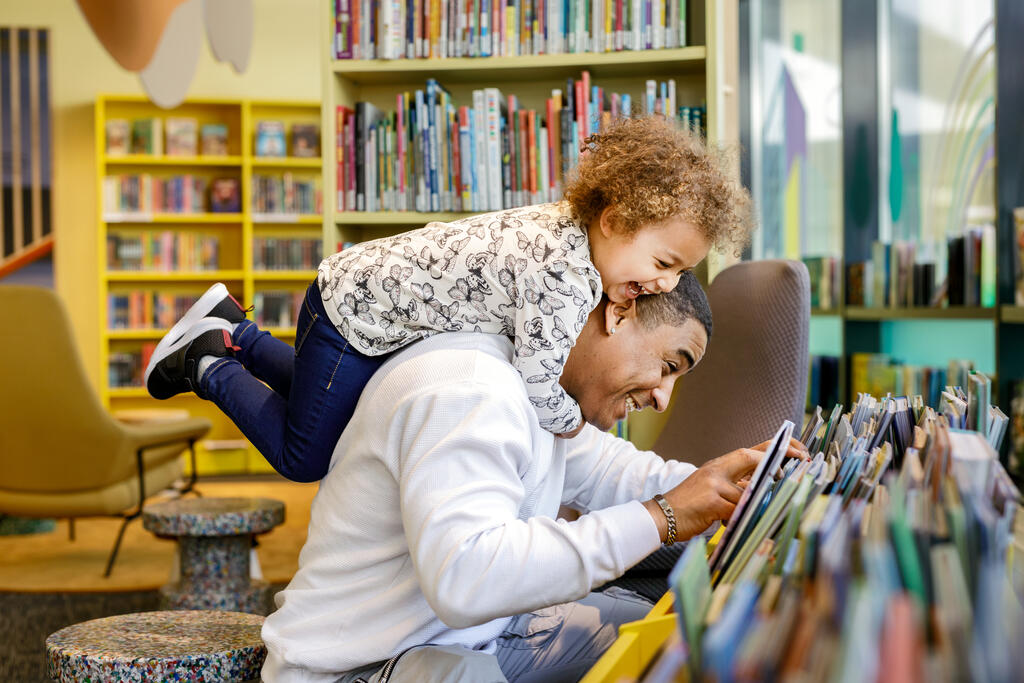  En man och ett barn väljer barnböcker i Kvarnbäckens biblioteks barnområde. Barnet har klättrat på mans rygg.