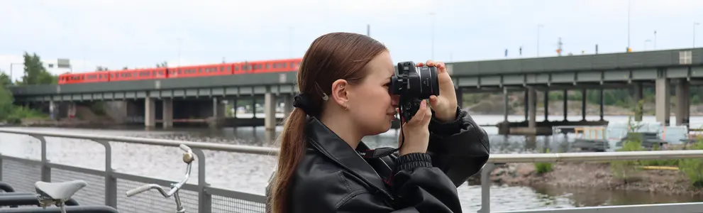 Anna Oikkonen kuvaa järjestelmäkameralla. Taustalla silta, jolla metro.