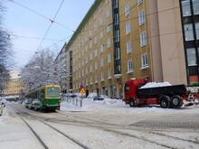Raitiovaunu seisoo lumisella kadulla. Asuintalojen vieressä on pysäköityjä autoja ja lunta pois vievä kuorma-auto.