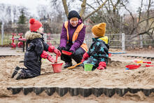 Kaksi päiväkoti-ikäistä lasta leikkii hiekkalaatikossa aikuisen kanssa kevätpäivänä.