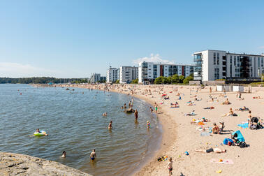 Ihmiset oleskelevat hiekkarannalla, taustalla kerrostaloja.